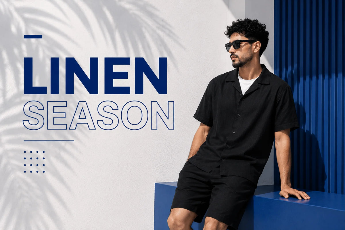 A man wearing a black linen short-sleeve button-up shirt and matching black shorts sits on a blue bench against a white and blue wall, with the text 'LINEN SEASON' on the left.